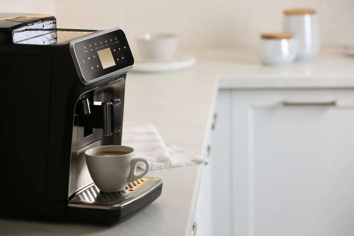 Une Tasse De Café Est Posée Sur Le Plateau D'une Machine à Café Automatique Noire Sur Un Comptoir De Cuisine, Avec Des Pots Et Des Plats En Arrière Plan.
