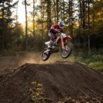 Un pilote de motocross en tenue complète saute un monticule de terre sur une piste forestière, avec la lumière du soleil filtrant à travers les arbres en arrière-plan.