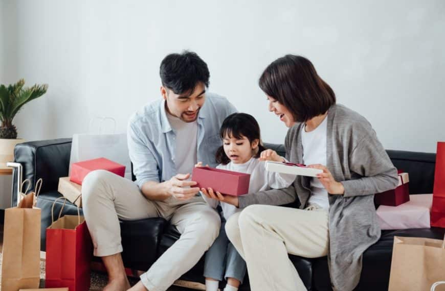 Une Famille De Trois Personnes Est Assise Sur Un Canapé, Ouvrant Ensemble Une Boîte Cadeau, Entourée De Sacs De Courses.