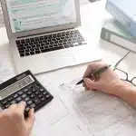 Une Personne Remplit Des Formulaires Fiscaux Avec Un Stylo, Utilise Une Calculatrice Et Référence Des Documents Sur Un Ordinateur Portable ; Des Classeurs Verts Et Une Plante En Arrière Plan.