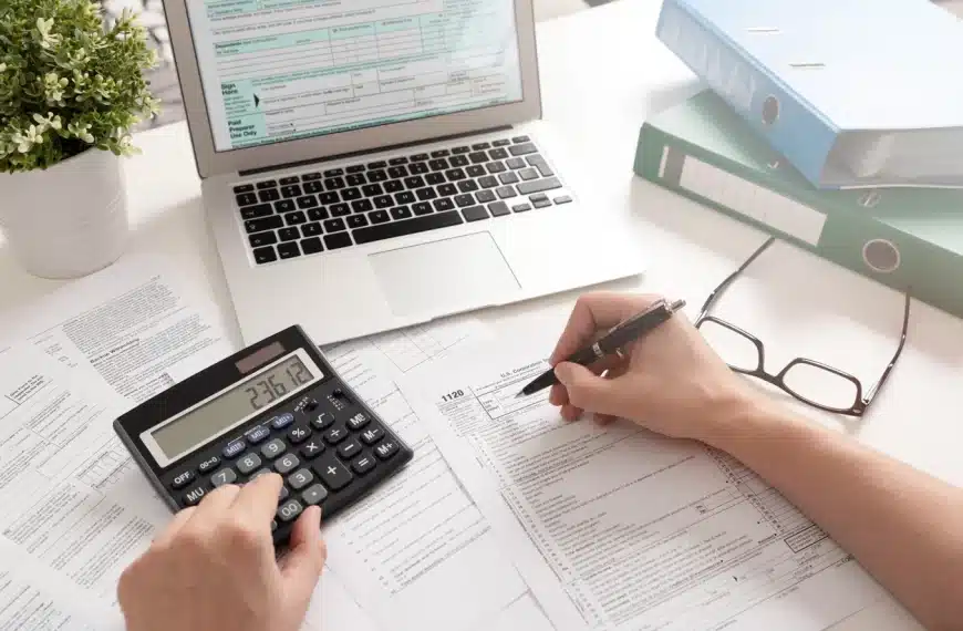 Une Personne Remplit Des Formulaires Fiscaux Avec Un Stylo, Utilise Une Calculatrice Et Référence Des Documents Sur Un Ordinateur Portable ; Des Classeurs Verts Et Une Plante En Arrière Plan.