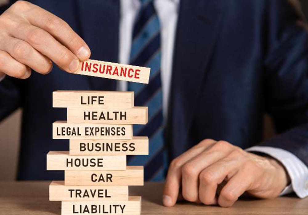 Une Personne En Costume Place Un Bloc De Bois étiqueté "assurance" Au Dessus D'une Pile De Blocs étiquetés Avec Des Catégories Telles Que Vie, Santé, Maison, Voiture, Voyage Et Responsabilité.