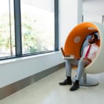 Un Homme En Tenue De Travail Est Assis Dans Un Fauteuil Moderne Près D'une Fenêtre, Utilisant Un Ordinateur Portable Et Portant Un Casque De Réalité Virtuelle.