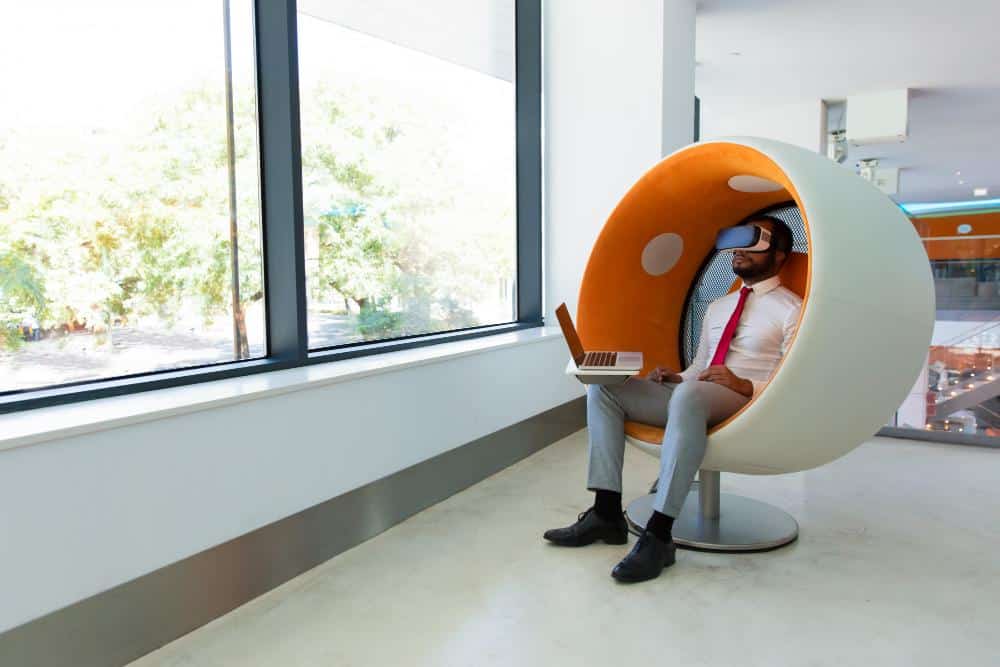 Un Homme En Tenue De Travail Est Assis Dans Un Fauteuil Moderne Près D'une Fenêtre, Utilisant Un Ordinateur Portable Et Portant Un Casque De Réalité Virtuelle.