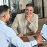 Deux Hommes Sont Assis à Un Bureau Et Discutent D'un Ordinateur Portable Affichant Des Graphiques Financiers. L'un D'eux Montre L'écran Tandis Que L'autre écoute En Souriant.