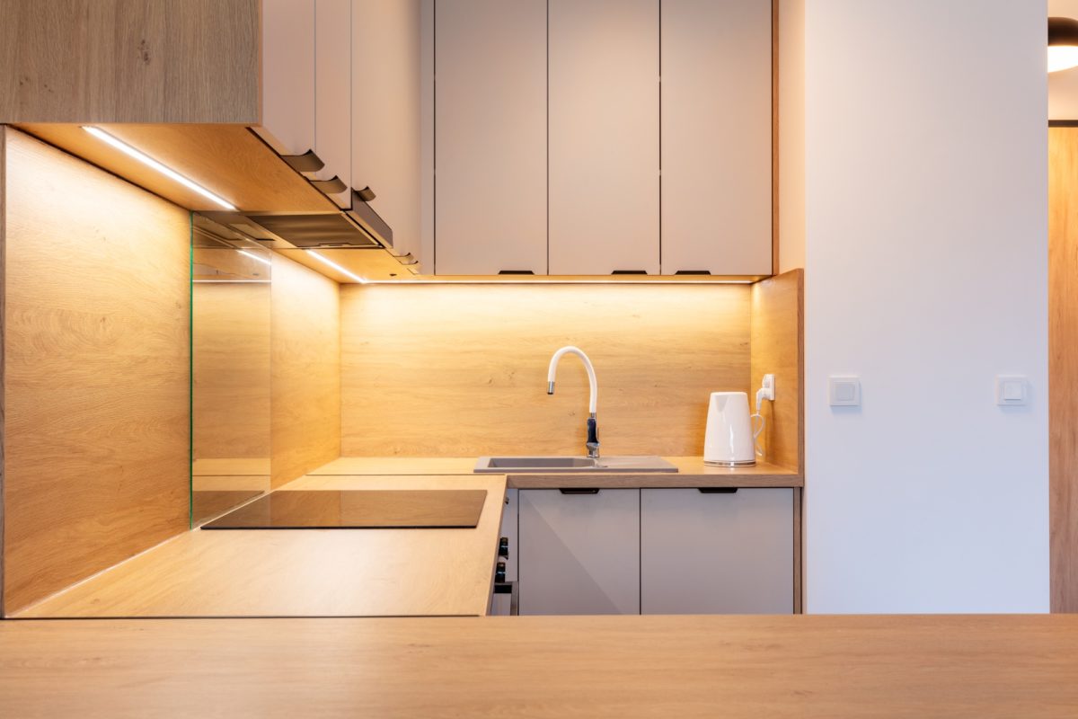 Cuisine Moderne Avec Des Meubles En Bois Et Beige, Un éclairage Sous L'armoire, Une Table De Cuisson électrique, Un évier Et Une Bouilloire électrique Blanche Sur Le Comptoir.