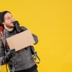 Un Homme Portant Un Sac à Dos Et Des Gants Tient Une Pancarte En Carton Vierge Et Semble Préoccupé Sur Un Fond Jaune.