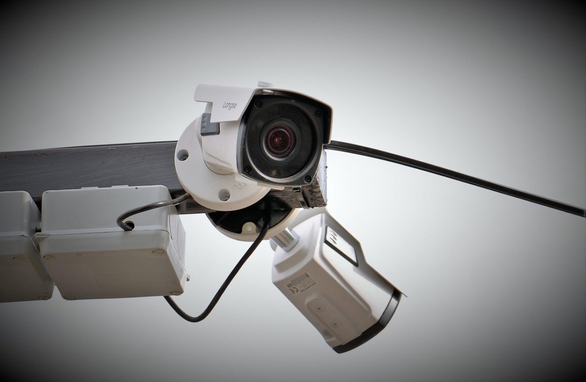 Vue Rapprochée De Deux Caméras De Surveillance Montées Sur Un Poteau Métallique Avec Des Câbles Visibles, Sur Un Fond Clair Et Simple.