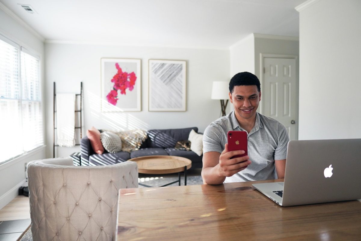 Un Homme Est Assis à Une Table En Bois, Utilisant Un Ordinateur Portable Et Tenant Un Smartphone Rouge, Avec Un Canapé Et Des Objets D'art Décoratifs Visibles à L'arrière Plan.
