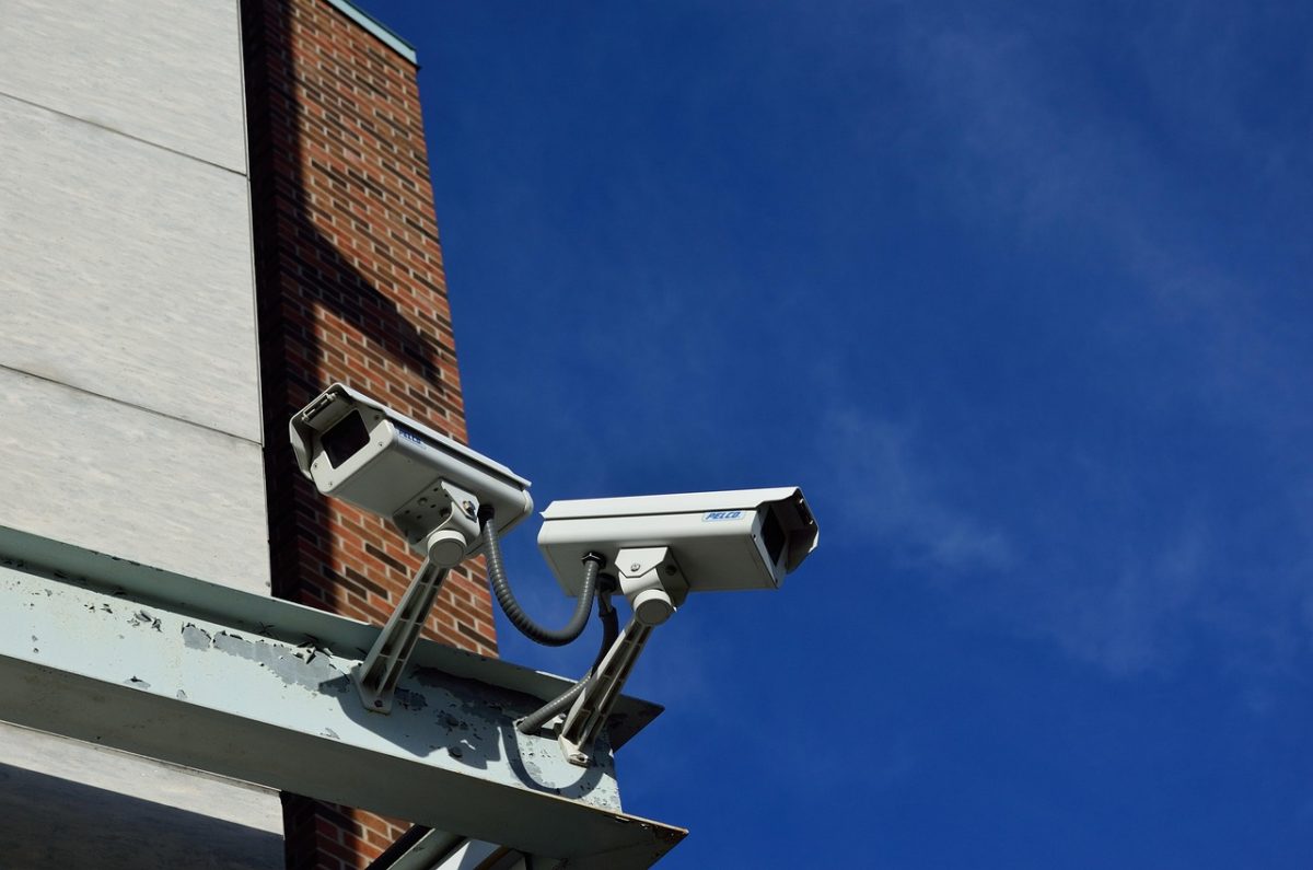 Deux Caméras De Sécurité Montées Sur Une Poutre Métallique à L'extérieur D'un Bâtiment Aux Murs De Briques Et De Béton, Orientées Dans Des Directions Légèrement Différentes Par Rapport à Un Ciel Bleu Clair.