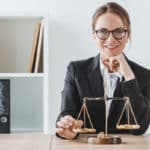 Une Femme En Tailleur Est Assise à Un Bureau Avec Une échelle De Justice Et Sourit à La Caméra. Des étagères Contenant Des Livres Et Des Dossiers Se Trouvent à L'arrière Plan.