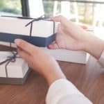 Une Personne Tenant Un Paquet Cadeau Emballé Avec Un Ruban Bleu à Une Table, Avec D'autres Paquets Cadeaux En Arrière Plan.