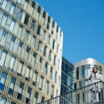 Un Homme En Costume Se Tient Sur Un Escalier Extérieur Et Parle Au Téléphone, Avec En Arrière Plan Des Immeubles De Bureaux Modernes En Verre Et Un Ciel Bleu Clair.