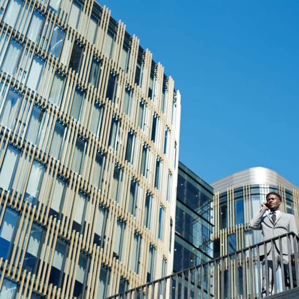 Un Homme En Costume Se Tient Sur Un Escalier Extérieur Et Parle Au Téléphone, Avec En Arrière Plan Des Immeubles De Bureaux Modernes En Verre Et Un Ciel Bleu Clair.
