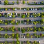 Vue Aérienne D'un Parking Avec Plusieurs Rangées De Voitures Garées Et Des Arbres Verts Régulièrement Espacés.