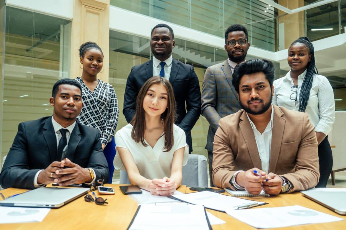 Sept Personnes Habillées Professionnellement Posent Pour Une Photo De Groupe à Une Table De Conférence Avec Des Ordinateurs Portables, Des Documents Et Des Stylos Dans Un Bureau Moderne.