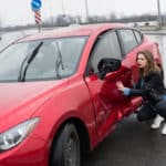 Une Femme Est Agenouillée à Côté D'une Voiture Rouge Dont La Portière Est Endommagée Et Le Rétroviseur Latéral Manquant, Sur Une Route Mouillée.