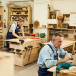 Deux Hommes Dans Un Atelier De Menuiserie ; L'un Travaillant Sur Un Ordinateur Portable à Une Table Au Premier Plan, L'autre Mesurant Du Bois à Un établi à L'arrière Plan.