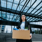 Une Femme En Tenue De Travail Se Tient à L'extérieur D'un Immeuble De Bureaux Moderne, Tenant Une Boîte En Carton Contenant Des Fournitures De Bureau, Ce Qui Laisse Supposer Qu'elle Quitte Son Emploi.