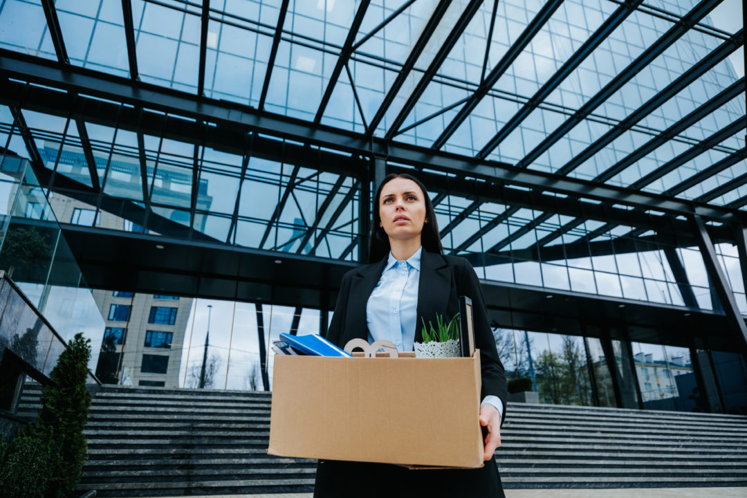 Une Femme En Tenue De Travail Se Tient à L'extérieur D'un Immeuble De Bureaux Moderne, Tenant Une Boîte En Carton Contenant Des Fournitures De Bureau, Ce Qui Laisse Supposer Qu'elle Quitte Son Emploi.