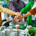 Cinq Personnes Empilent Leurs Mains Dans Un Geste De Travail D'équipe Au Dessus D'une Table Où Se Trouvent Des Modèles Verts, Des éoliennes Et Des Tasses De Café, Suggérant Un Projet De Développement Durable Collaboratif.