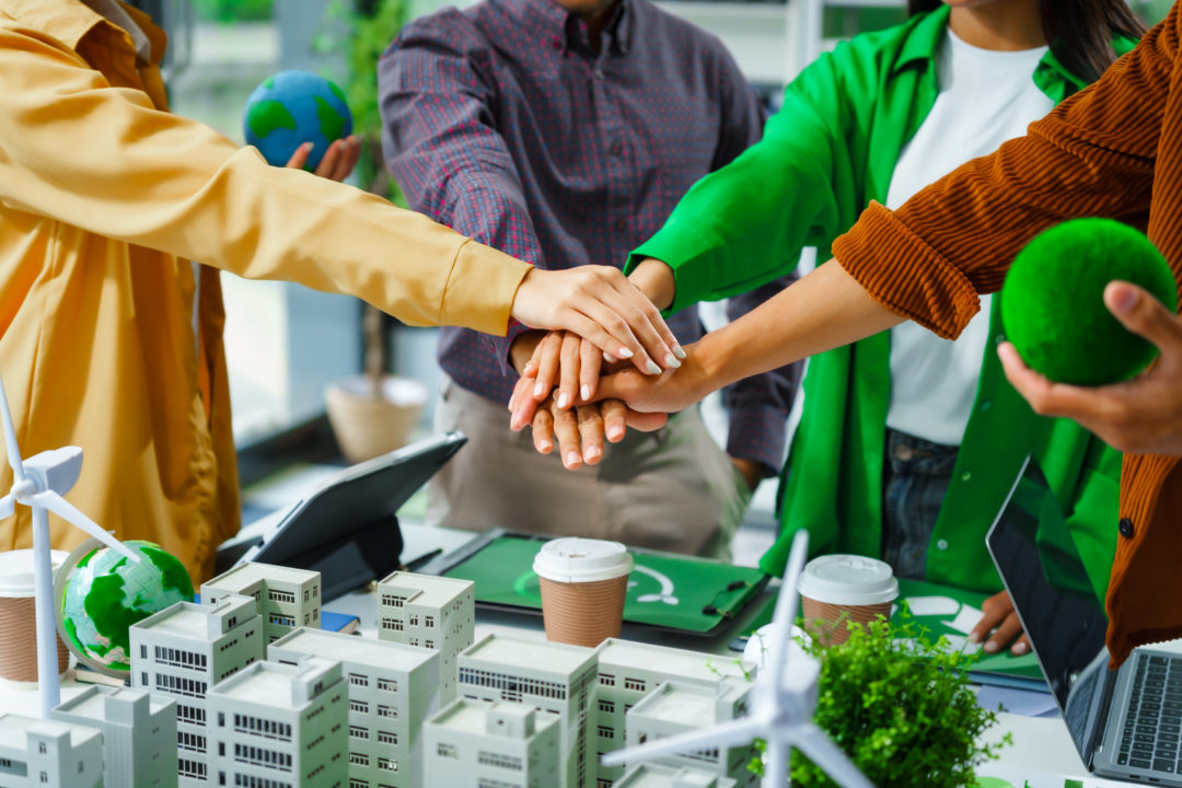 Cinq Personnes Empilent Leurs Mains Dans Un Geste De Travail D'équipe Au Dessus D'une Table Où Se Trouvent Des Modèles Verts, Des éoliennes Et Des Tasses De Café, Suggérant Un Projet De Développement Durable Collaboratif.