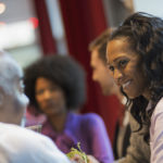 Quatre Personnes Sont Assises Ensemble Et Discutent à L'intérieur ; L'accent Est Mis Sur Une Femme Qui Sourit à Un Homme De L'autre Côté De La Table, Deux Autres Personnes étant Floues à L'arrière Plan.