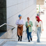 Trois Femmes Portant Un Foulard Montent Des Escaliers Extérieurs à Côté D'un Bâtiment Moderne, En Discutant Et En Souriant ; L'une D'elles Tient Une Tasse De Café.