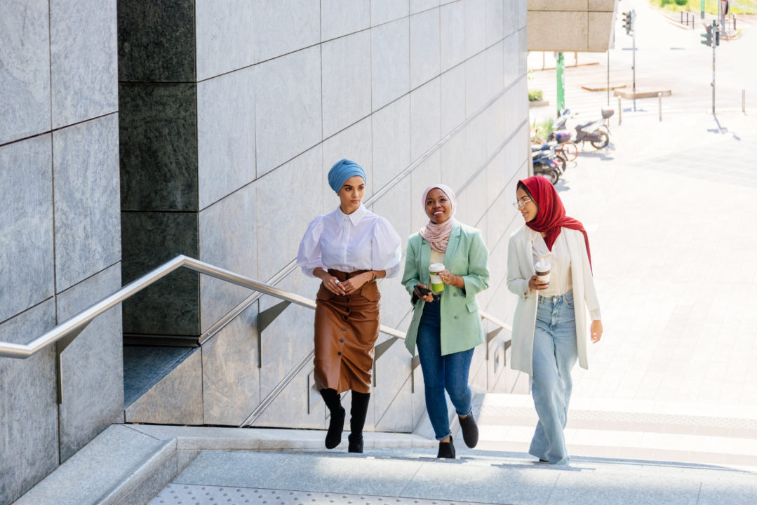 Trois Femmes Portant Un Foulard Montent Des Escaliers Extérieurs à Côté D'un Bâtiment Moderne, En Discutant Et En Souriant ; L'une D'elles Tient Une Tasse De Café.