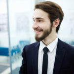 Un Jeune Homme En Costume Cravate Se Tient à L'intérieur, Souriant Et Regardant Sur Le Côté, Avec Des Meubles De Bureau Et Des Personnes Floues à L'arrière Plan.
