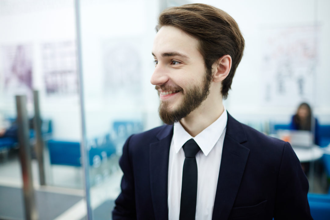 Un Jeune Homme En Costume Cravate Se Tient à L'intérieur, Souriant Et Regardant Sur Le Côté, Avec Des Meubles De Bureau Et Des Personnes Floues à L'arrière Plan.