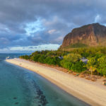 Vue Aérienne D'une Plage De Sable Fin Bordée De Parasols Et De Chaises Longues, Bordée D'une Eau Turquoise Et D'une Végétation Dense, Avec Une Grande Montagne En Arrière Plan Sous Un Ciel Nuageux.