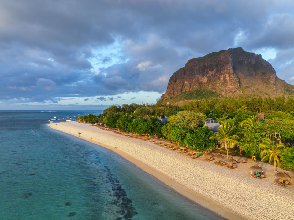 Vue Aérienne D'une Plage De Sable Fin Bordée De Parasols Et De Chaises Longues, Bordée D'une Eau Turquoise Et D'une Végétation Dense, Avec Une Grande Montagne En Arrière Plan Sous Un Ciel Nuageux.