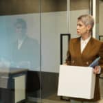 Une Personne Aux Cheveux Courts, Vêtue D'un Costume Marron, Tient Une Boîte Et Des Dossiers En Quittant Un Bureau Par Une Porte Vitrée.