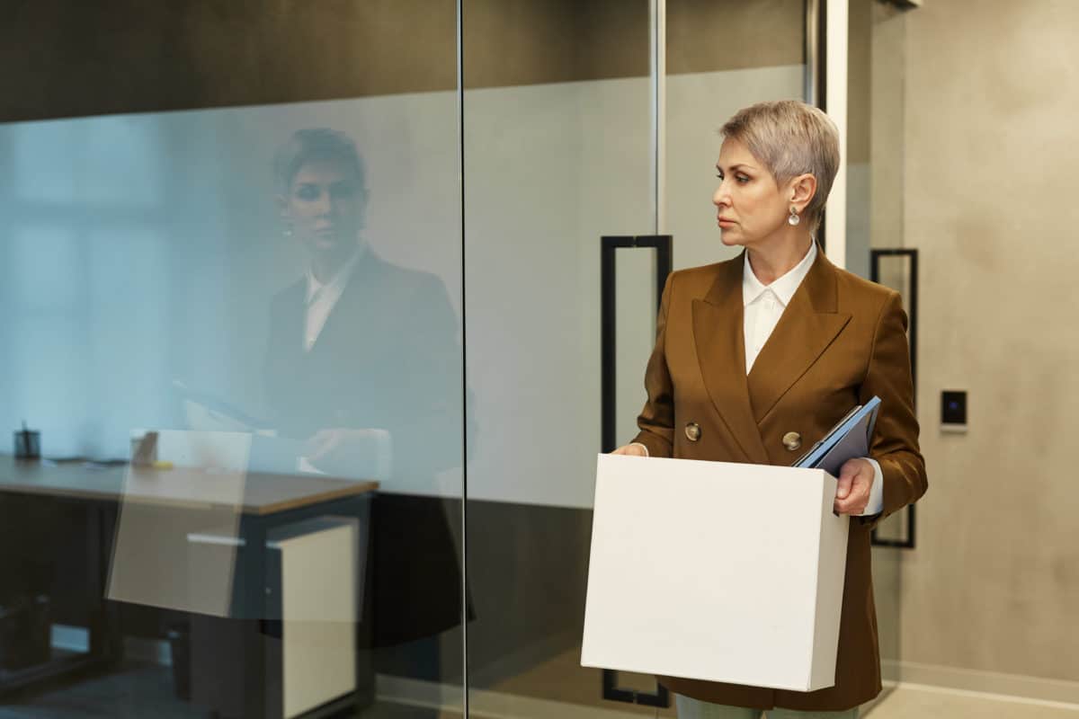 Une Personne Aux Cheveux Courts, Vêtue D'un Costume Marron, Tient Une Boîte Et Des Dossiers En Quittant Un Bureau Par Une Porte Vitrée.