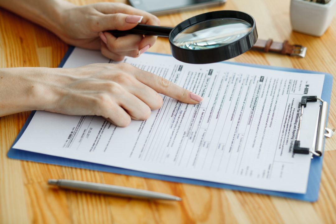 Une Personne Tient Une Loupe Et Pointe Une Partie D'un Formulaire Sur Un Presse Papiers, Avec Un Stylo Et Un Smartphone Sur Un Bureau En Bois.