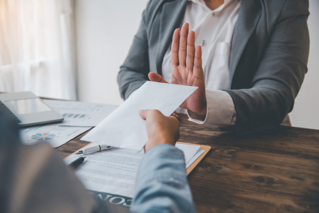 Une Personne En Costume Refuse Une Enveloppe Proposée Par Une Autre Personne En Face D'un Bureau Avec Des Documents Et Un Stylo.