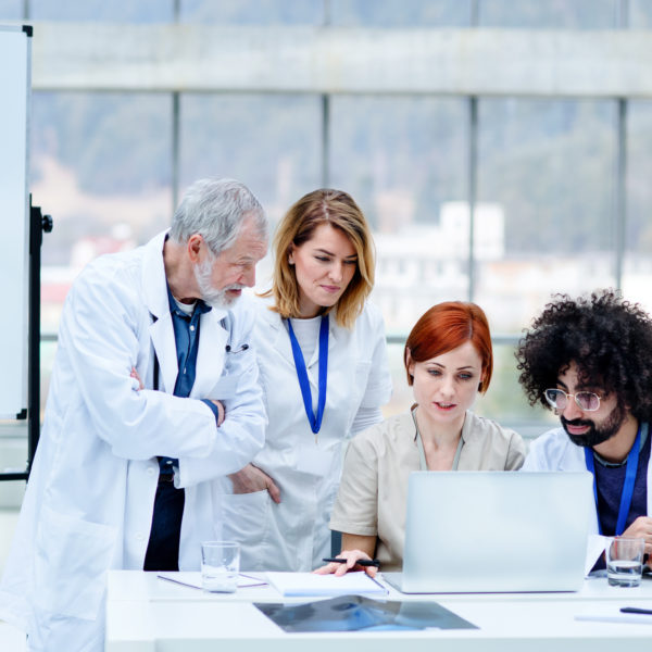 Dans Une Salle Moderne Et Lumineuse, Quatre Professionnels De La Santé Consultent Des Informations Sur Un Ordinateur Portable Installé à Un Bureau, Tandis Que Des Images Radiographiques S'affichent Sur Un écran à Proximité.