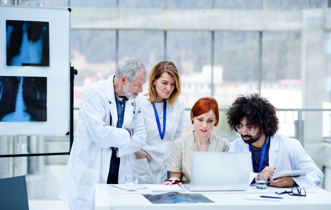 Dans Une Salle Moderne Et Lumineuse, Quatre Professionnels De La Santé Consultent Des Informations Sur Un Ordinateur Portable Installé à Un Bureau, Tandis Que Des Images Radiographiques S'affichent Sur Un écran à Proximité.