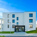 Immeuble Moderne De Trois étages Avec Un Extérieur Blanc, De Grandes Fenêtres Et Une Pelouse Sous Un Ciel Bleu Clair.