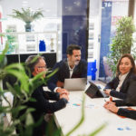 Quatre Personnes En Tenue De Travail Sont Assises Autour D'une Table De Conférence Avec Des Ordinateurs Portables Et Des Documents. Elles Participent à Une Réunion Dans Un Bureau Moderne Avec Des Plantes Et Des Murs En Verre.