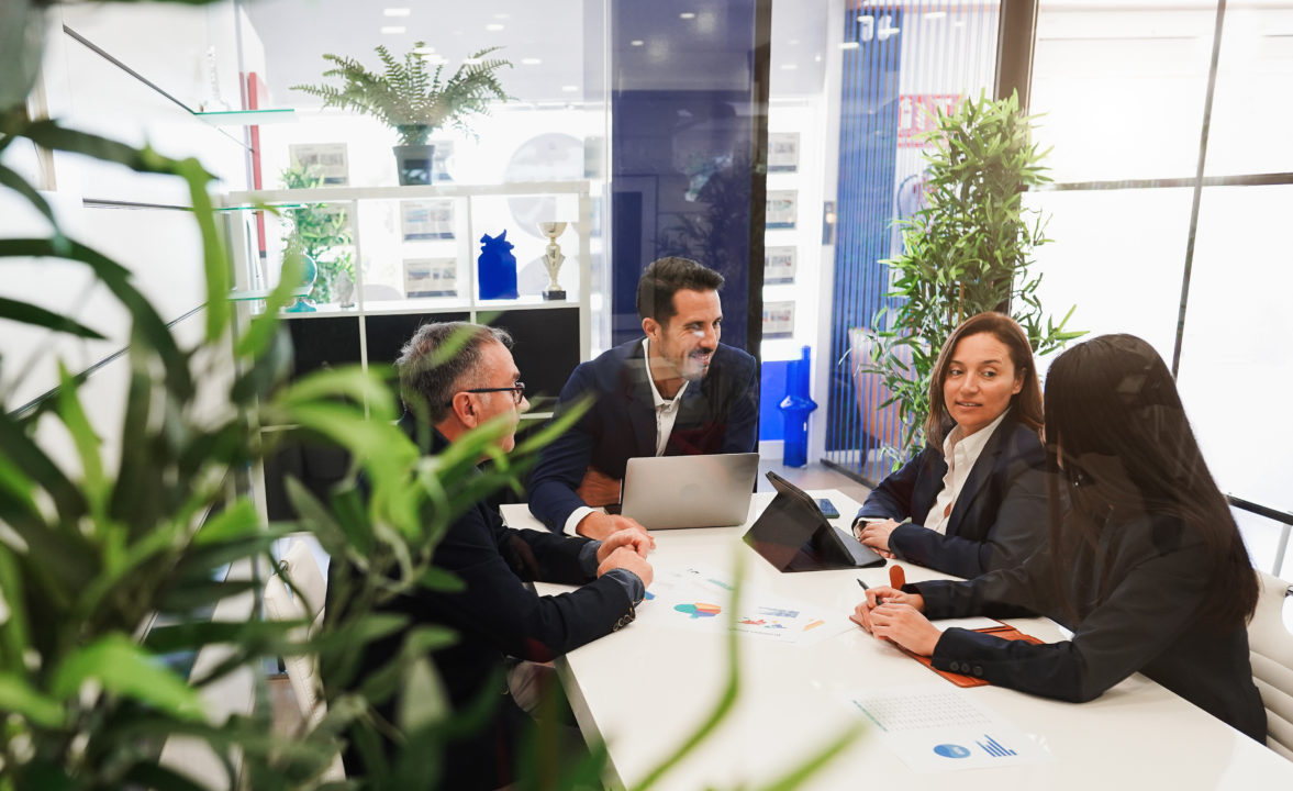 Quatre Personnes En Tenue De Travail Sont Assises Autour D'une Table De Conférence Avec Des Ordinateurs Portables Et Des Documents. Elles Participent à Une Réunion Dans Un Bureau Moderne Avec Des Plantes Et Des Murs En Verre.