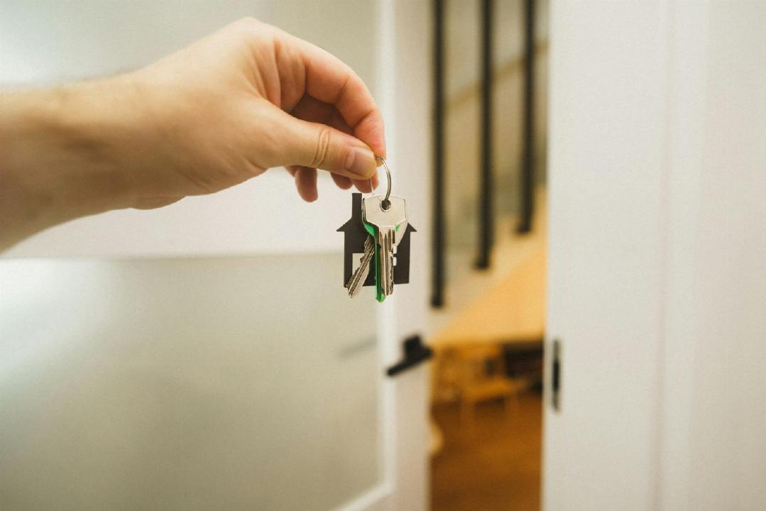 Une Main Tient Un Trousseau De Clés Sur Un Porte Clés Devant Une Porte Ouverte, Avec Un Intérieur Flou Visible à L'arrière Plan.