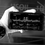 Une Personne Tenant Un Smartphone Et Affichant Un Graphique Financier, Avec En Arrière Plan Un Graphique Mural Montrant Des Tendances à La Hausse.