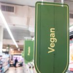 Un Panneau Vert Portant L'inscription "vegan" Est Accroché Dans Une Allée D'épicerie, Avec Des étagères De Produits Et Un Autre Panneau Visible à L'arrière Plan.
