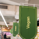 Un Panneau Vert De L'épicerie Portant Le Mot "vegan" Est Suspendu Au Dessus D'une Allée, Indiquant La Section Réservée Aux Produits Végétaliens.