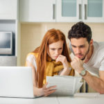 Une Femme Et Un Homme Dans Une Cuisine Regardent Des Documents Avec Des Expressions Préoccupées Tout En Utilisant Un Ordinateur Portable.