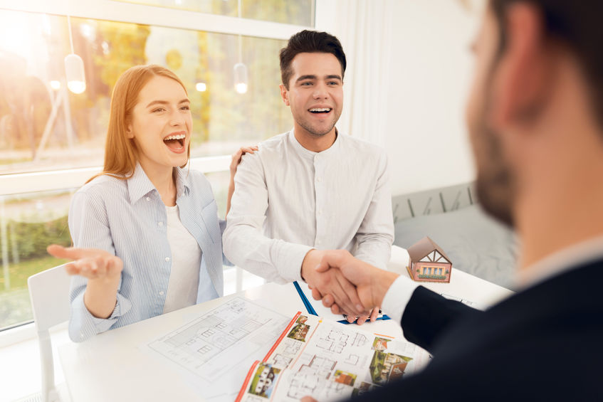 Un Couple Souriant Est Assis à Une Table Avec Des Plans De Maison Et Une Maison Miniature, Tout En Serrant La Main D'une Autre Personne, Ce Qui Suggère Une Réunion Sur L'immobilier Ou L'achat D'une Maison.