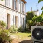 Une Pompe à Chaleur Est Installée Dans Le Jardin D'une Maison En Pierre Aux Volets Blancs, Entourée De Plantes Et De Verdure Par Une Journée Ensoleillée.
