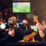 Trois Personnes Sont Assises à Une Table Dans Un Bar, Tenant Des Boissons Et Regardant Un Match De Football Sur Un écran De Télévision En Arrière Plan.